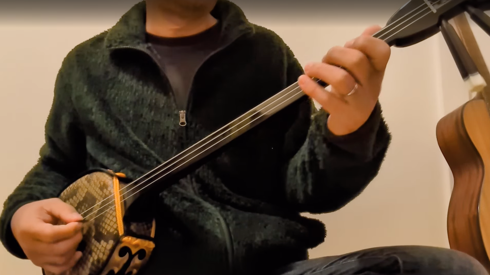 三線の練習をする男性。背景にはアコースティックギター