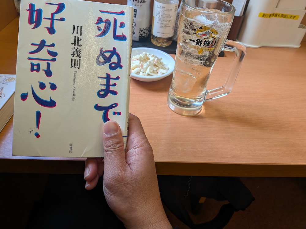 「死ぬまで好奇心！（川北義則著）」表紙と背景にハイボールとマカロニサラダ