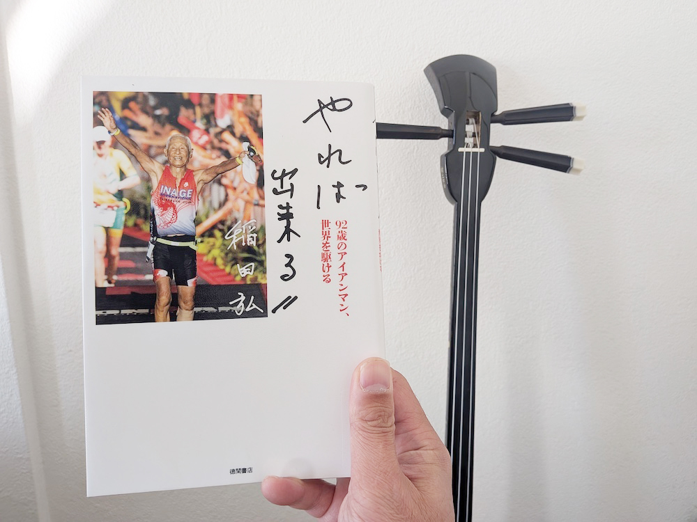 「やれば出来る!!92歳のアイアンマン、世界を駆ける(稲田弘著)」の表紙と背景に三線
